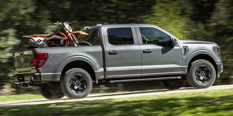 A gray 2024 Ford F-150 with a red motor cross bike in the bed, driving in a park near Cheboygan, MI