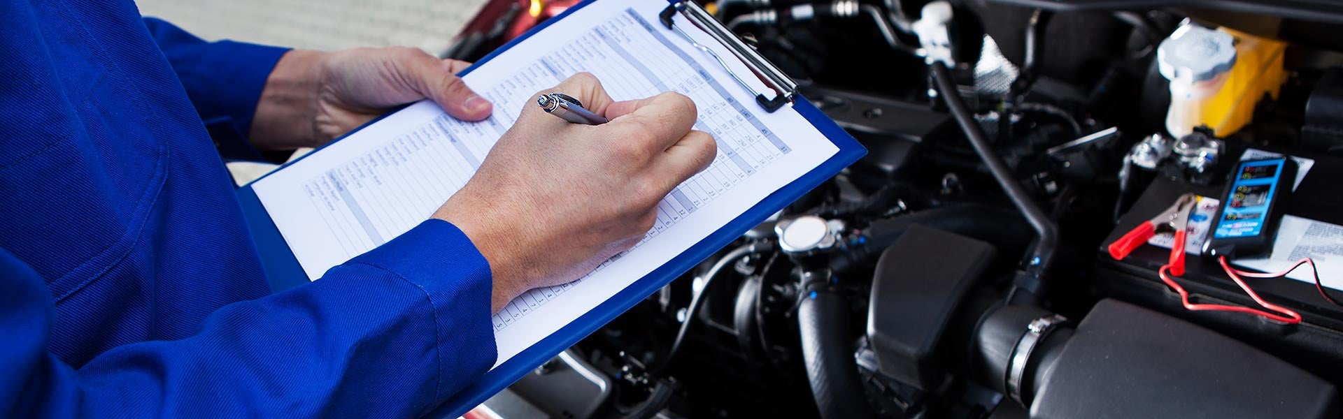 Mechanic writing on a checklist clipboard over an open car engine bay